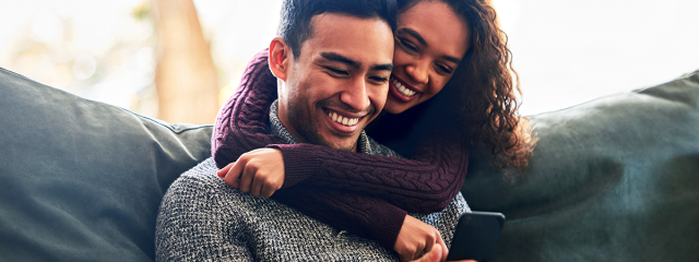 A couple at home smiling and looking at a phone with arms around each other