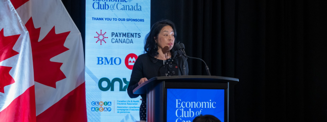 A shot from behind an audience of a Stephanie speaking at a dark podium, with a bright blue vertical banner behind.