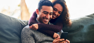 A couple at home smiling and looking at a phone with arms around each other